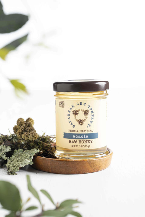 A jar of Acacia honey placed on a wooden surface with a label that includes the brand name and product name. There are also some acacia tree leaves and blossoms visible in the background.