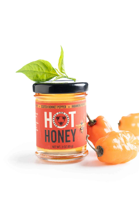 A jar of Hot Honey with a scotch bonnet pepper and a leaf garnish on top, placed next to a fresh habanero pepper.