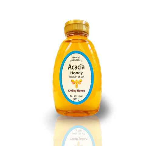 A jar of Acacia honey placed on a wooden surface with a label that includes the brand name and product name. There are also some acacia tree leaves and blossoms visible in the background.