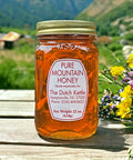 Jar of pure mountain honey with a label on a wooden surface, surrounded by flowers and mountains in the background.