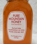 A jar of Pure Mountain Honey with a label that includes the brand name and contact information, placed on a table with a scenic background of mountains and flowers.