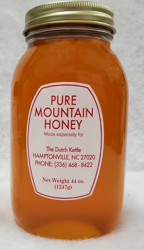 A jar of Pure Mountain Honey with a label that includes the brand name and contact information, placed on a table with a scenic background of mountains and flowers.