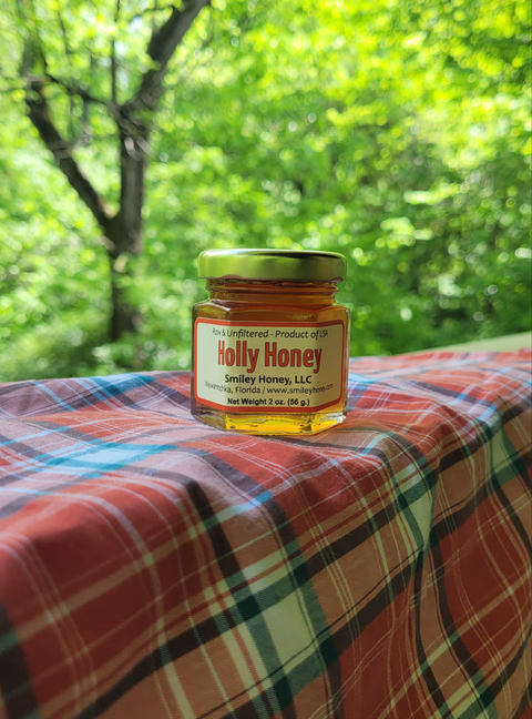 A jar of Holly Honey placed on a table with a red checkered tablecloth, with a background of greenery.