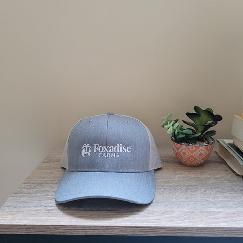 A gray snapback trucker hat with the 'Foxadise Farms' logo embroidered on the front, displayed on a wooden surface next to a small potted plant.