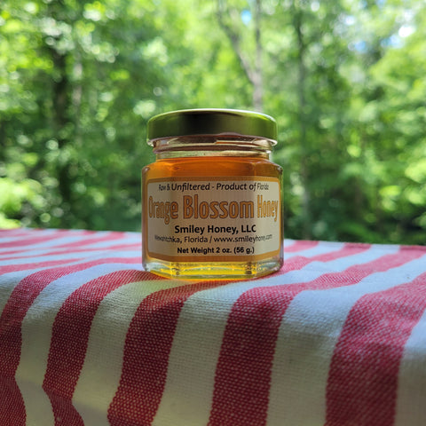 Jar of orange blossom honey from Smiley Honey, LLC on a red and white striped fabric with a blurred green forest background.