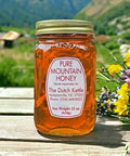 A jar of Pure Mountain Honey with a label that includes the brand name and contact information, placed on a table with a scenic background of mountains and flowers.