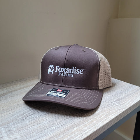 Brown and beige cap with 'Foxadise Farms' logo on a wooden surface.
