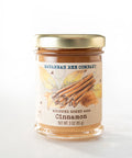 A jar of Savannah Bee Company's Whipped Honey with Cinnamon, showing a clear glass jar filled with light colored honey, sealed with a gold-colored lid and a label that includes an image of cinnamon sticks.