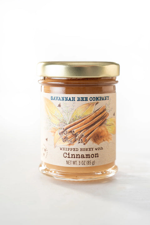 A jar of Savannah Bee Company's Whipped Honey with Cinnamon, showing a clear glass jar filled with light colored honey, sealed with a gold-colored lid and a label that includes an image of cinnamon sticks.